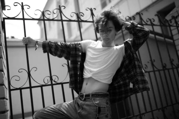 Stylish confident young guy wearing jeans and plaid shirt standing near wrought-iron fencing on city street on summer day, tidying curly wavy hair by hand. Fashion black and white photo