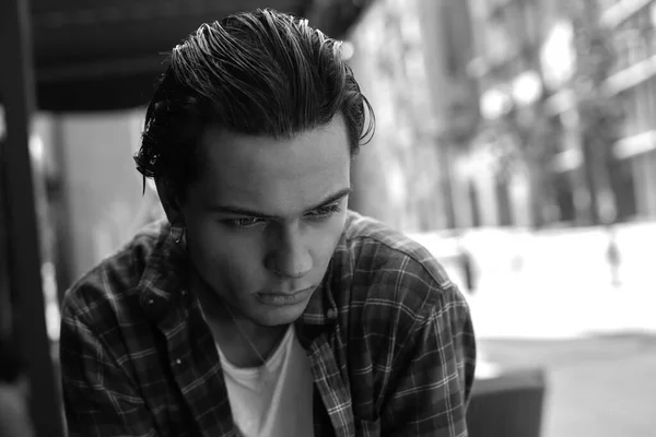 Closeup portrait in black and white of sad pensive dark-haired young guy sitting outdoors against blurred background on summer cityscape