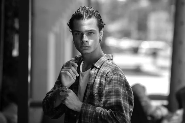 Black and white portrait of handsome dark-haired young guy in casual plaid shirt posing while walking along summer street of modern city, looking confidently at camera