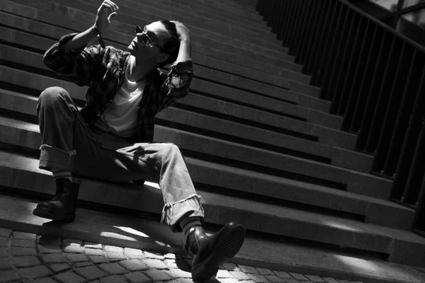 Stylish young guy wearing jeans and plaid shirt sitting on stone staircase on city street, confidently adjusting black curly hair. Modern urban lifestyle concept. Black and white photography