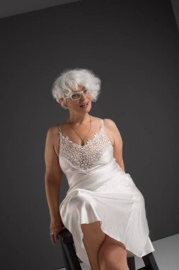 Gri arka planda, ahşap taburede neşeli bir şekilde gülümseyen beyaz ipek dantelli kaşkorse giymiş, kıvırcık bukleli gözlüklü, gümüş saçlı, yaşlı bir kadın.