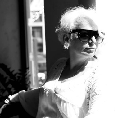 Sakin, rüya gibi, gri saçlı, açık renk elbiseli, güneşli, güneşli ve koyu güneş gözlüğüyle pencereden dışarı bakan yaşlı kadın. Siyah beyaz portre