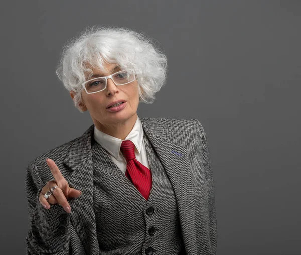 Confident stylish silver-haired mature businesswoman in strict formal suit and red tie posing on gray studio background, gesturing towards something with hand. Female boss giving instruction