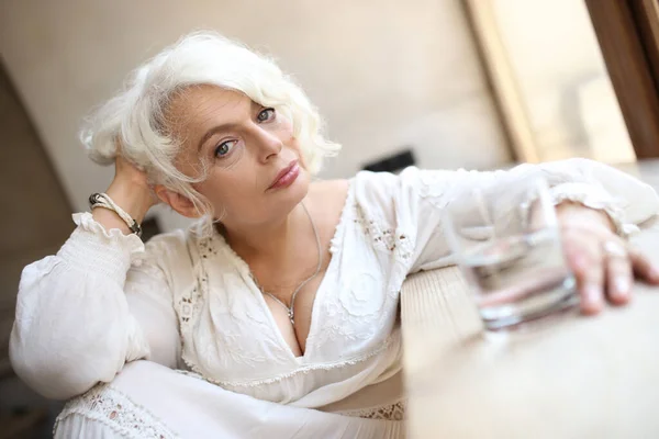 Serene attractive silver-haired elderly woman in light white dress sitting by window with glass of water leaning on wooden windowsill and supporting head with hand, calmly looking at camera