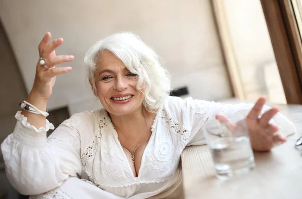 Cheerful attractive silver-haired elderly woman in light white dress sitting by window with glass of water leaning on wooden windowsill,  gesturing emotionally and laughing, looking at camera