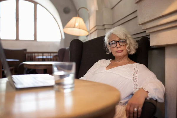 Portrait of stylish silver-haired aged woman in glasses and light white dress relaxing in comfortable easy chair while working on laptop standing on table
