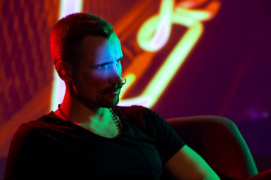 Rahat, şık, sakallı, gidon bıyıklı genç bir adam. Loş bir gece kulübünde arka planda, yumuşak koltukta oturuyor. Parlak neon ışıklarının altında.