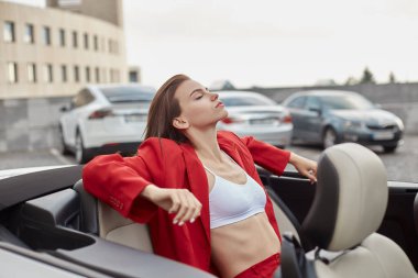 Beyaz bluz ve kırmızı takım elbiseli, rahat rahat oturan, açık havada park edilmiş, üstü açık arabadaki arka koltukta oturan, temiz yaz havasının tadını çıkaran, rahat, rahat ve rahat genç bir kız.