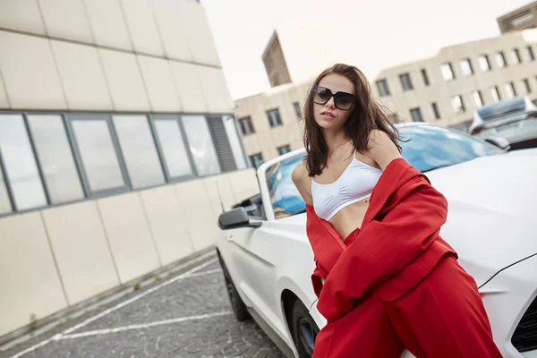 Portrait of confident relaxed young brunette in red pantsuit with blazer pulled off her shoulders and sunglasses standing leaning against hood of convertible car