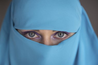 Mavi gözlü genç kadın turkuaz kumaş giyiyor, kameraya bakıyor..