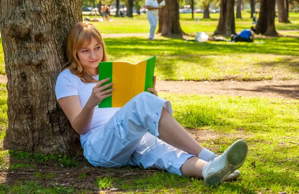 一个拿着一本书的女孩坐在公园的一棵树旁 — 图库照片
