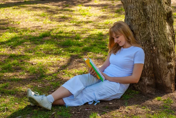 一个拿着一本书的女孩坐在公园的一棵树旁 — 图库照片
