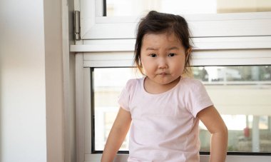 Genç kız bir pencerenin önünde oturuyor, pembe bir gömlek giyiyor. Üzgün görünüyor ve pencereden dışarı bakıyor.