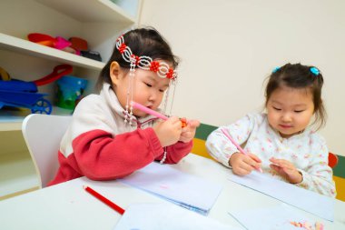 İki genç kız bir masada oturuyor, çiziyorlar ve yazıyorlar. Bir kız saç bandı takıyor ve diğeri beyaz gömlek giyiyor. İkisi de kalem kullanıyor ve eğleniyor gibi görünüyorlar.