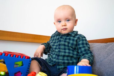 Bebek blok ve kovayla oynuyor. Bebek yerde oturuyor ve elinde bir oyuncak var.