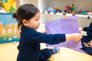 Genç kız yerde oturuyor ve elinde bir kağıt parçası var. O, kağıda bakıyor ve konsantre oluyor gibi görünüyor. Kız görevini yaparken odaklanma ve kararlılık kavramı