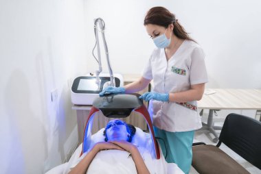 Bir kadın tıbbi bir ortamda bir hastayı tedavi ediyor. Hasta maske takıyor ve bir yatakta yatıyor. Kadın beyaz laboratuvar önlüğü giyiyor ve hastayı tedavi etmek için bir cihaz kullanıyor.