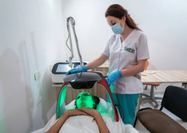 Kadın, tıbbi bir ortamda bir hastayı tedavi ediyor. Hasta maske takıyor ve bir yatakta yatıyor. Kadın beyaz laboratuvar önlüğü giyiyor ve hastayı tedavi etmek için bir cihaz kullanıyor.