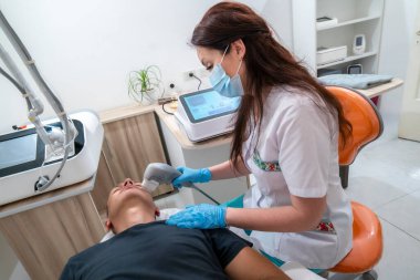 Beyaz laboratuvar önlüklü bir kadın bir adamın kafasını cihazla tedavi ediyor. Adam siyah bir gömlek giyiyor.