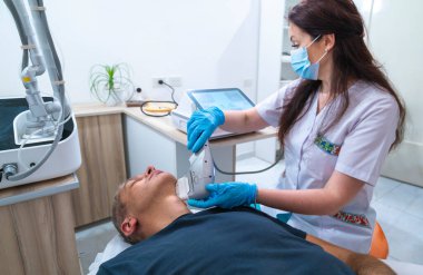 Beyaz laboratuvar önlüklü bir kadın bir adamın kafasını cihazla tedavi ediyor. Adam siyah bir gömlek giyiyor.