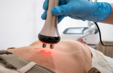 Bir adam karnında kırmızı ışıkla masaj yaptırıyor. Adam mavi eldiven giyiyor.