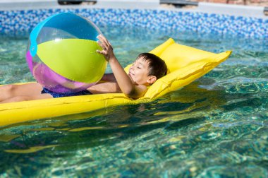 Genç bir çocuk elinde mavi ve yeşil bir plaj topuyla havuzda yüzüyor. Sahne neşeli ve eğlenceli, çocuğun suda geçirdiği zamanın tadını çıkarıyor.
