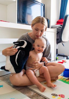 Bir kadın kucağında bebek, yerde oturuyor. Bebek gülümsüyor ve kadın bebeğe bakıyor. Yakınlarda bir penguen oyuncağı var.