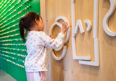 Genç bir kız büyük bir harf bloğuyla bir duvara resim çiziyor. Harfler 