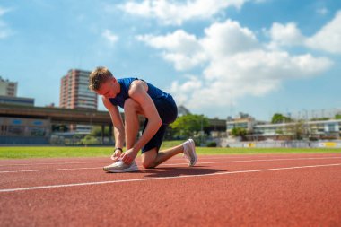 Bir adam pistte koşmaya hazırlanıyor. Mavi bir gömlek ve siyah bir şort giyiyor.