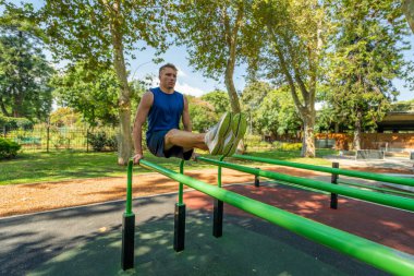 Adamın biri parktaki bankta amuda kalkıyor. Bankta yeşil ve metal bir çerçeve var. Park ağaçlar ve otlarla dolu. Huzurlu bir ortam yaratıyor.