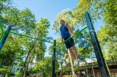 Parktaki bir barda barfiks çeken bir adam var. Park ağaçlarla dolu ve huzurlu bir atmosferi var.