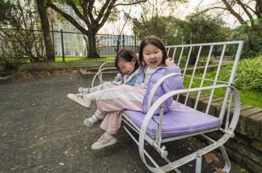 Parkta bir bankta oturan iki genç kız. İçlerinden biri mor bir ceket giyiyor.