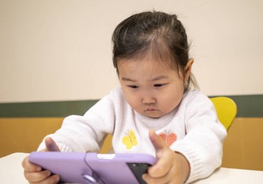 Genç bir kız tablette oyun oynuyor. Beyaz bir kazak giyiyor ve üzerinde kelebek var.