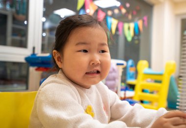 Genç bir kız yüzünde bir gülümsemeyle bir masada oturuyor. Beyaz bir kazak giyiyor ve gözleri kapalı. Oda renkli afişlerle süslenmiş durumda. Atmosfer neşeli görünüyor.