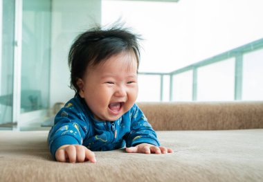 Bir bebek kanepede yatarken gülümsüyor ve gülüyor. Bebek mavi bir gömlek giyiyor.