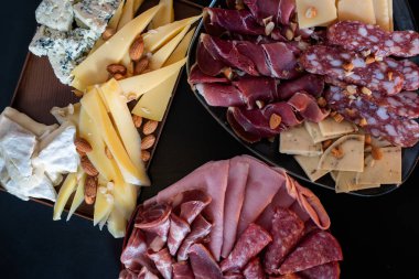 Siyah arka planda çeşitli et ve peynir tabakları sergileniyor. Etler arasında dilimlenmiş jambon, salam ve muhtemelen diğer et türleri yer alıyor. Peynirler arka planda.