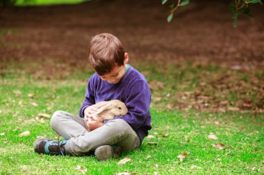 Kucağında bir tavşanla çimlerin üzerinde oturan bir çocuk. Tavşan küçük ve beyaz