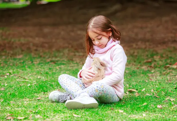 Bir kız çimlerin üzerinde elinde bir tavşan tutuyor. Pembe bir kapüşonlu giyiyor. Tavşan küçük ve tüylü.