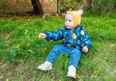 Küçük bir çocuk çimlerin üzerinde mavi ceket ve kot pantolon giyiyor. Ceketin üzerinde sarı bir şapka var.