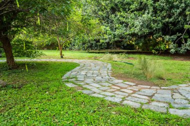 Taşlardan yapılmış bir patika bir parkta. Yol çimenler ve ağaçlarla çevrili. Arka planda birkaç ağaç var ve patikanın yakınında bir bank var.
