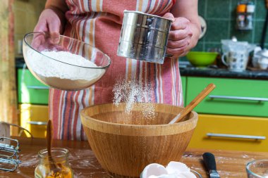 Mutfak masasının yanında dururken ahşap kasenin üzerinde ev yapımı hamur işi için un toplayan önlüklü kadının elleri..
