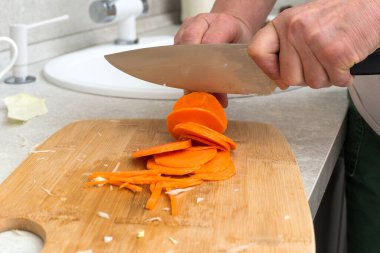 Kadın eli, mutfak masasında tahta bir tahtanın üzerinde bıçakla havuç kesiyor. Pişirmeden önce taze havuç keser..