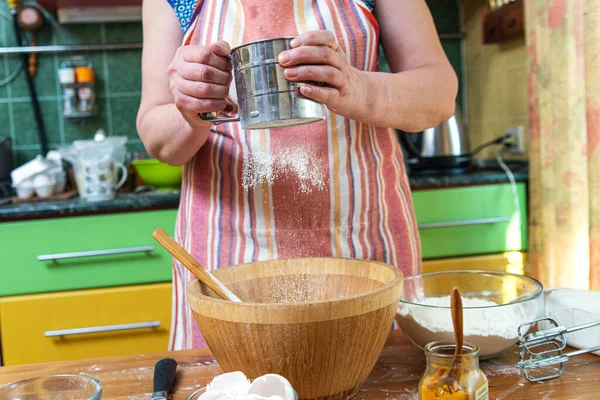 站在厨房桌旁用围裙的女人的手在木碗上筛粉做自制的糕点 — 图库照片