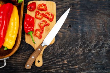 Kesme tahtasında bıçakla taze çiğ tatlı biber. Doğranmış biber parçaları pişmeye hazır. Koyu ahşap masa, üst görünüm. Metin için boşluk..