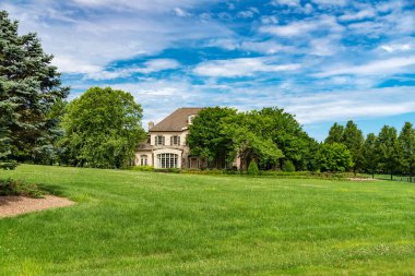 büyük, lüks, tuğla gibi iki katlı büyük bir aile evi peyzajı ve kocaman yeşil bir çimenliği var. Mavi bulutlu gökyüzünün altında güneşli bir yaz günü manzarası.