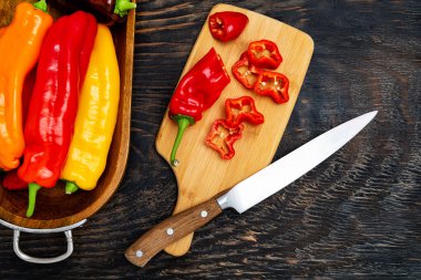 Kesme tahtasında bıçakla taze çiğ tatlı biber. Doğranmış biber parçaları pişmeye hazır. Koyu ahşap masa, üst manzara..
