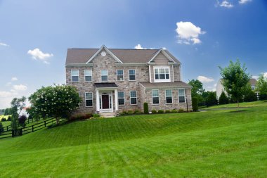 Büyük, lüks, peyzajlı tuğladan bir ev ve kocaman yeşil bir çimen. Mavi bulutlu gökyüzünün altında güneşli bir yaz günü manzarası.