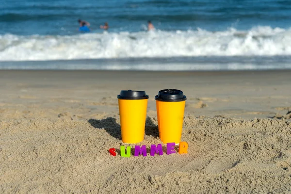 Two paper cups with coffee on the sand by the ocean. The inscription SUMMER in colored letters.