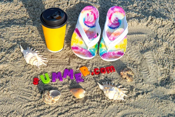 beach slippers, a paper cup with coffee and sea shells on the beach sand with the inscription SUMMER.
