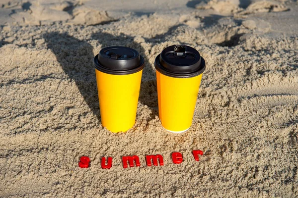 yellow paper cups with coffee on the sand in the sunset rays of the sun. The inscription SUMMER in red letters.